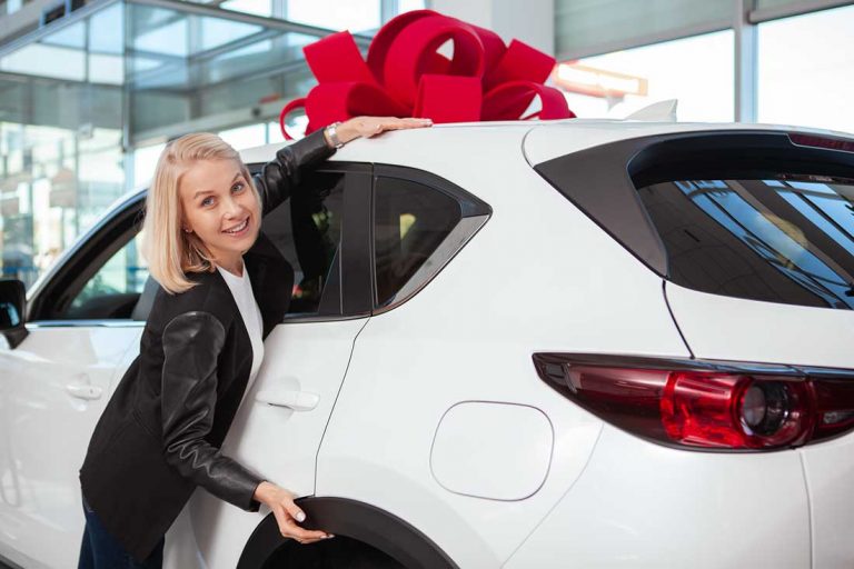 happy-beautiful-woman-hugging-her-new-car-with-red-bow-on-the-roof-cheerful-female-driver-receiving-new-car-as-a-gift-at-dealership